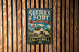 A vintage metal sign for Sutter's Fort State Historic Park in California, featuring a wooden fence and a cow in the background.