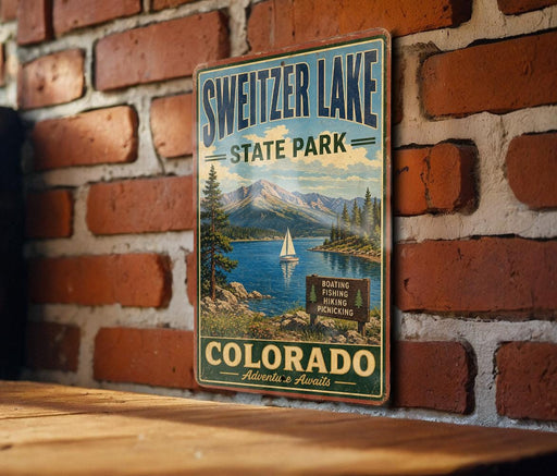 A vintage-style metal sign for Sweitzer Lake State Park in Colorado, featuring a scenic landscape with a lake, mountains, and a sailboat.