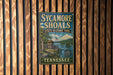 A vintage-style metal sign for Sycamore Shoals State Historic Park in Tennessee, featuring a scenic illustration of a lake and a historic building.