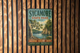 A vintage-style sign for Sycamore State Park in Ohio, featuring a scenic landscape with a lake and trees.