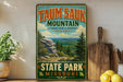 A vintage-style sign for Taun sauk Mountain, the highest point in Missouri, with a scenic mountain landscape and pine trees.