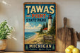 A vintage-style metal sign for Tawas Point State Park in Michigan, featuring a lighthouse and a beach scene.