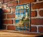 A vintage metal sign for Tawas Point State Park, featuring a lighthouse and beach scene, is displayed on a brick wall.