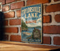 A vintage-style metal sign for Taylorville Lake State Park in Kentucky, featuring a scenic illustration of a lake, boats, and a sign welcoming visitors to explore, relax, and enjoy the natural surroundings.