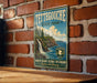 A vintage-style metal sign for Tettegouche State Park, featuring a scenic landscape with a waterfall and mountains, mounted on a brick wall.