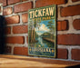 A vintage-style metal sign for Tick Faw State Park in Louisiana, featuring a scenic illustration of a park with a wooden walkway, a pond, and a bridge.