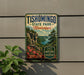 A vintage-style metal sign for Tishomingo State Park in Mississippi, featuring a scenic landscape with a river, cliffs, and flowers.