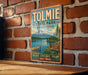 A vintage-style metal sign for Tolmie State Park in Washington, featuring a scenic mountain landscape and a wooden dock.