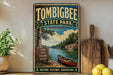A vintage-style metal sign for Tombbee State Park in Mississippi, featuring a scenic image of a river, pine trees, and a canoe.