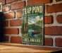 A metal sign with a painting of a red canoe on a lake, surrounded by trees, is mounted on a brick wall.