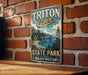 A vintage metal sign for Triton Cove State Park in Washington, featuring a scenic landscape with a beach, mountains, and pine trees.