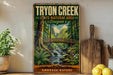 A vintage-style metal sign for "Tryon Creek State Natural Area Oregon" featuring a scenic illustration of a bridge over a river surrounded by lush greenery.