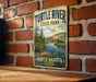 A vintage-style metal sign for Turtle River State Park in North Dakota, featuring a scenic illustration of a river and forest landscape.
