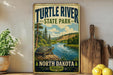 A vintage-style sign for Turtle River State Park in North Dakota, featuring a scenic illustration of a turtle on a rock by a river surrounded by pine trees.