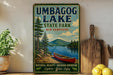 A vintage-style sign for Umbagog Lake State Park in New Hampshire, featuring a scenic image of a red canoe on a lake surrounded by pine trees and mountains.