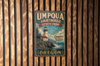 A vintage-style metal sign for Umpqua Lighthouse State Park, featuring a lighthouse and a coastal scene, is displayed on a wooden fence.