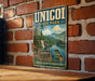 A vintage metal sign for Unicoi State Park, featuring a scenic landscape with a waterfall and mountains, mounted on a brick wall.