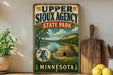 A vintage-style metal sign for Upper Sioux Agency State Park in Minnesota, featuring a scenic illustration of the park and its surroundings.