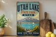 A vintage-style sign for Utah Lake State Park, featuring a scenic illustration of a lake, mountains, and a sailboat, along with the text "UTAH LAKE STATE PARK UTAH" and "BOATING, FISHING, HIKING, PICNICKING Adventure Awaits!"