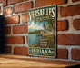 A vintage metal sign for Versailles State Park, featuring a scenic illustration of a lake and a lighthouse, is displayed on a brick wall.