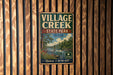 A vintage-style metal sign for Village Creek State Park in Texas, featuring a scenic landscape with a river, trees, and a cloudy sky.