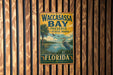A vintage-style metal sign for Waccassas Bay Preserve in Florida, featuring a bird and a palm tree, is displayed on a wooden wall.