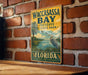 A vintage-style metal sign for Waccasass Bay Preserve State Park in Florida, featuring a bird and palm trees, is displayed on a brick wall.