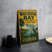 A vintage-style metal sign for Waccasass Bay Preserve in Florida, featuring a bird and palm trees, is displayed on a gray concrete surface.