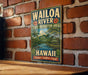 A vintage-style metal sign for the Wailoa River State Recreation Area in Hawaii, featuring a tropical landscape with palm trees, a river, and a beach.