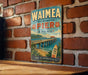 A vintage-style sign for Waimea Pier in Hawaii, featuring a wooden pier with a bridge, palm trees, and a surfboard, against a brick wall.