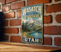 A vintage-style metal sign for Wasatch Mountain State Park in Utah, featuring a scenic mountain landscape with pine trees and a lake.