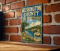A vintage-style metal sign for Washington County, Illinois, featuring a scenic landscape with a river, trees, and flowers.