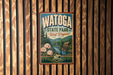A vintage-style poster for Watoga State Park in West Virginia, featuring a scenic landscape with a river, pine trees, and a wooden cabin.