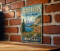 A vintage metal sign for Wayfarers State Park in Montana, featuring a scenic mountain landscape with a lake and pine trees.