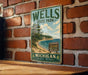 A vintage metal sign for Wells State Park, featuring a scenic landscape with a lake, mountains, and forests.