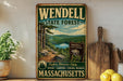 A vintage metal sign for Wendell State Forest in Massachusetts, featuring a scenic landscape with pine trees, a lake, and mountains.
