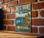 A vintage metal sign for West Shore State Park in Montana, featuring a scenic landscape with mountains, water, and pine trees.