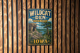 A vintage-style metal sign for Wildcat Den State Park in Iowa, featuring a scenic landscape with a rocky cliff, trees, and a river.