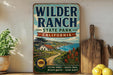 A vintage-style sign for Wilder Ranch State Park in California, featuring a scenic landscape with a house, trees, and a path leading to the ocean.