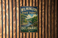 A vintage metal sign for Wilmington State Parks in Delaware, featuring a scenic landscape with a river and bridge.