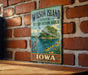 A vintage-style metal sign for Wilson Island, Iowa, featuring a scenic landscape with a lake, a bird, and flowers.