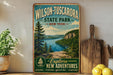 A vintage-style metal sign for Wilson-Tuscador State Park in New York, featuring a scenic landscape with a lake and mountains.