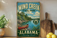 A vintage-style metal sign for Wind Creek State Park in Alabama, featuring a scenic image of a canoe on a river surrounded by pine trees.