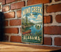 A vintage metal sign for Wind Creek State Park in Alabama, featuring a scenic image of a canoe on a lake surrounded by pine trees.