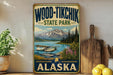 A vintage-style sign for Wood-Tikik State Park in Alaska, featuring a scenic image of a lake, mountains, and a seaplane.