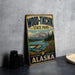 A vintage-style sign for Wood-Tikik State Park in Alaska, featuring a scenic landscape with a lake, mountains, and a plane.