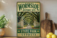 A vintage-style sign for Wormsloe Historic Site in State Park, Georgia, featuring a path lined with trees and a stone archway.