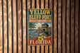 A vintage-style metal sign for the historic Bluff Fort in Florida, featuring a palm tree and a building, is displayed on a wooden wall.