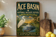 A vintage-style sign for Ace Basin National Wildlife Refuge in South Carolina, featuring an illustration of a white egret and a crocodile in a tropical setting.