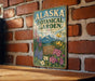 A vintage-style metal sign for the Alaska Botanical Garden, featuring illustrations of flowers and plants, is displayed on a brick wall.